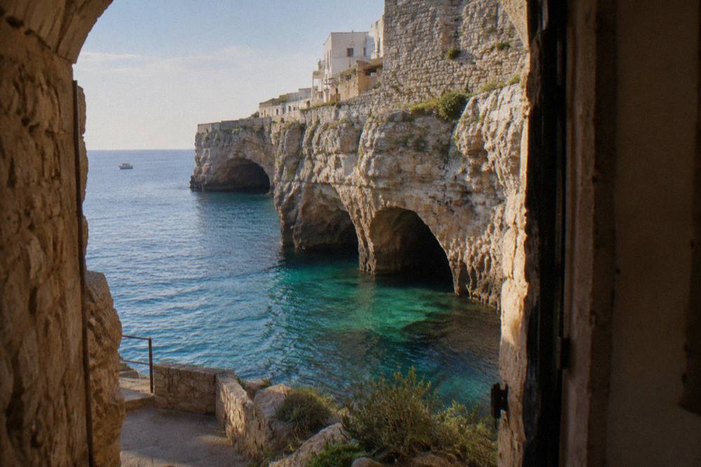 Sea cave openings at the base of tall limestone cliffs with turquoise water and a small boat far off