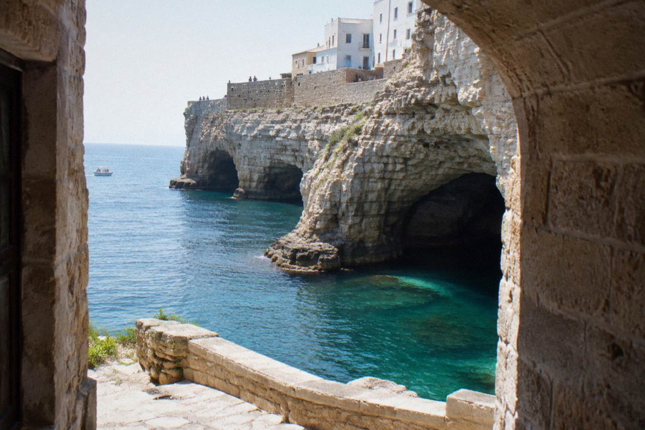 Sea cave openings at the base of tall limestone cliffs with turquoise water and a small boat far off