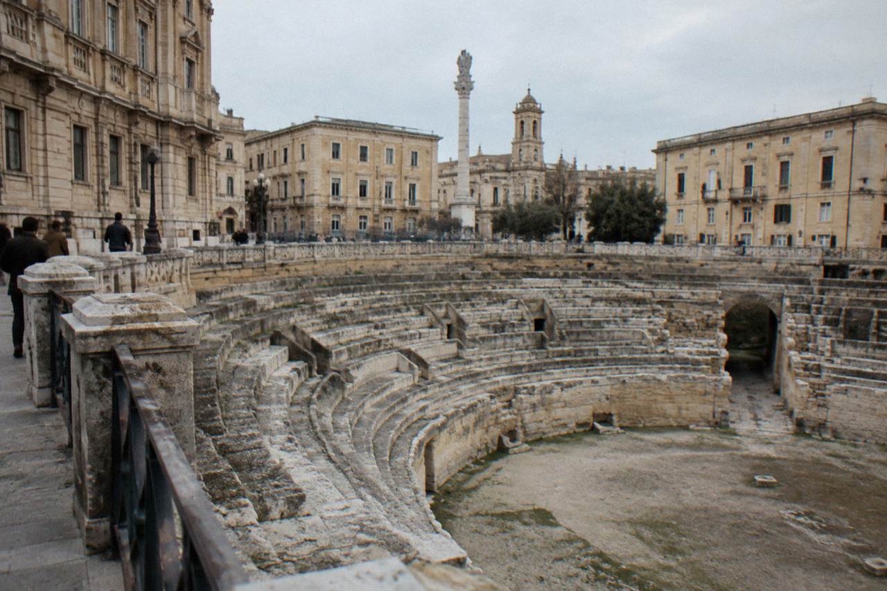 Roman amphitheatre half excavated in a city square surrounded by palazzi in afternoon light