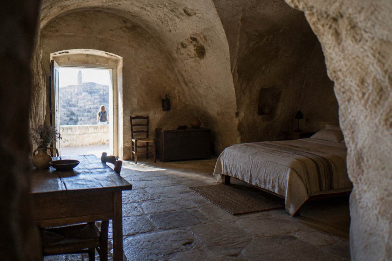 Inside a restored cave dwelling with an arched rock ceiling, a simple bed and a wooden table