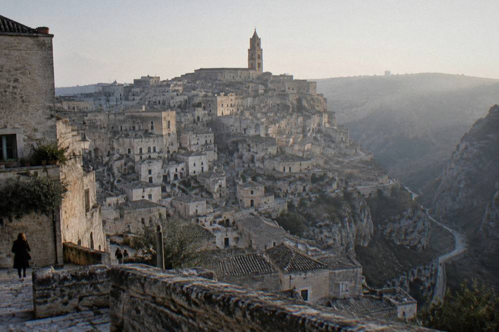 Tiers of honey-coloured cave dwellings stacked on the side of a deep ravine below a cathedral bell tower