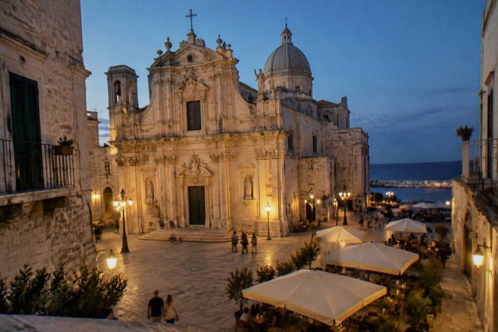 Baroque cathedral facade in warm pale stone on a small square with a dome behind and cafe tables