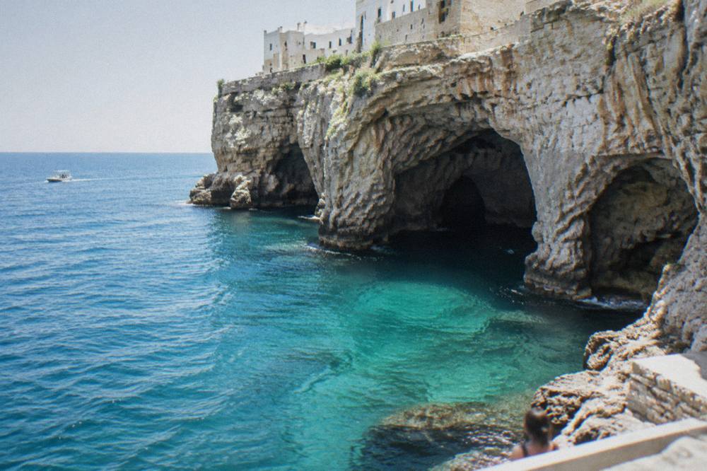 Sea cave openings at the base of tall limestone cliffs with turquoise water and a small boat far off