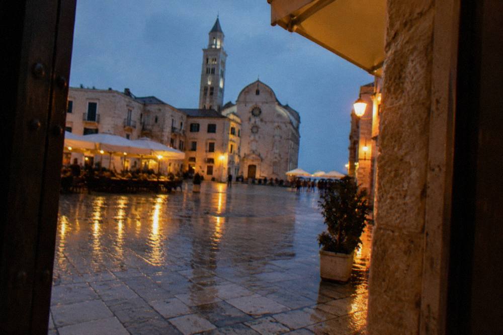 Rainy evening in a pale stone square with wet paving reflecting warm cafe lights and distant umbrellas