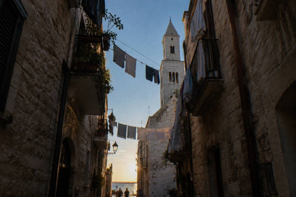 Looking up between close walls at a strip of blue sky crossed by washing lines and a bell tower