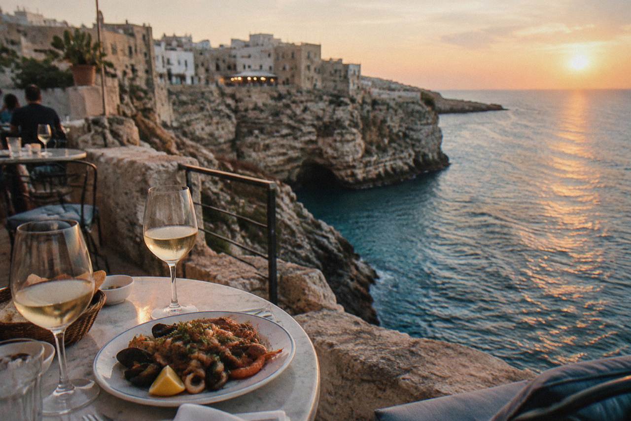 Small round tables on a cliff-edge terrace with glasses and a plate of seafood above blue water