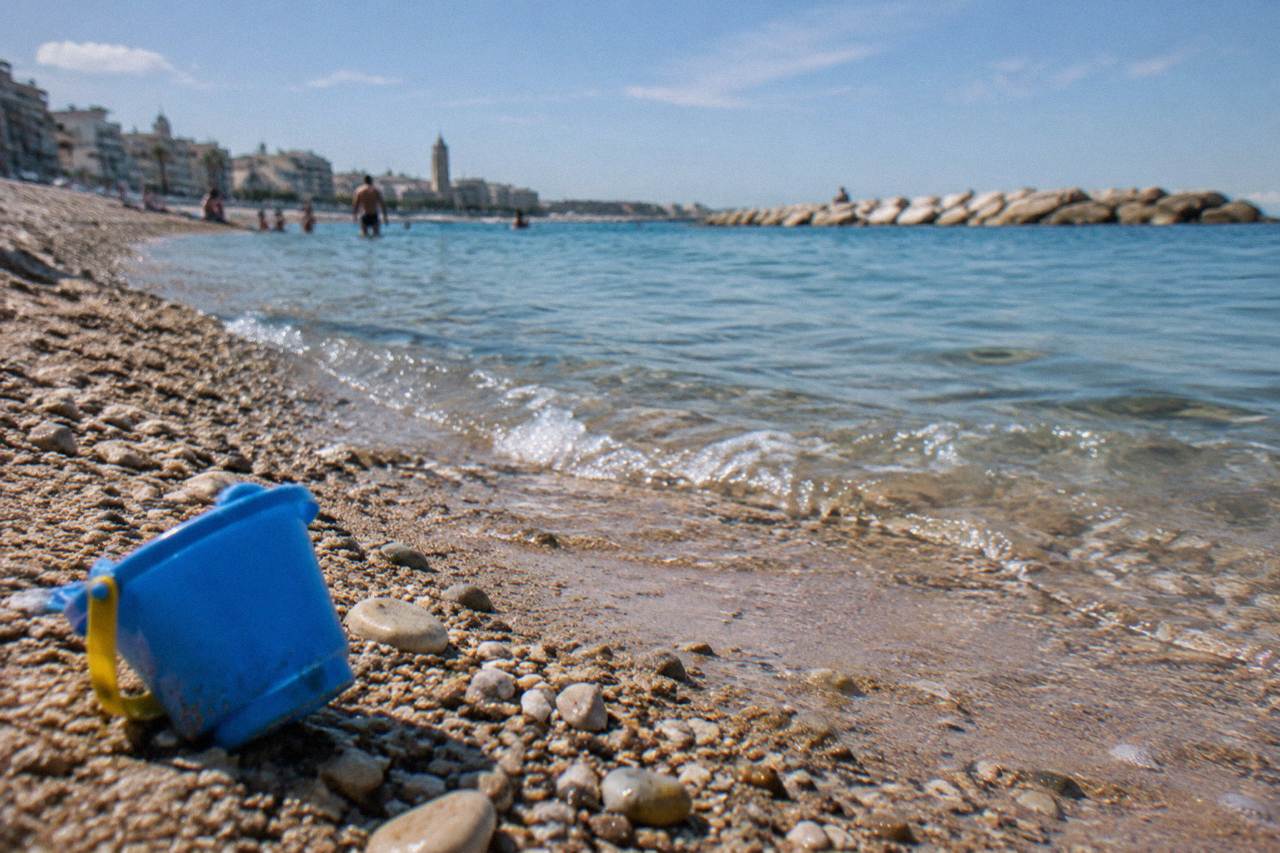 Pebbles and coarse sand at the water's edge with small clear waves and a plastic bucket left on the sand