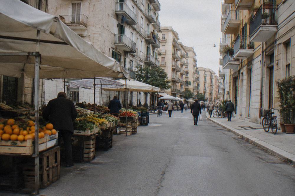 Residential street of apartment blocks with fruit and vegetable stalls under plain awnings and distant shoppers