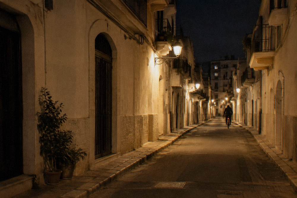 Quiet side street at night with warm lamps, a distant cyclist and pale stucco facades