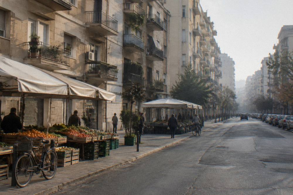Residential street of apartment blocks with fruit and vegetable stalls under plain awnings and distant shoppers