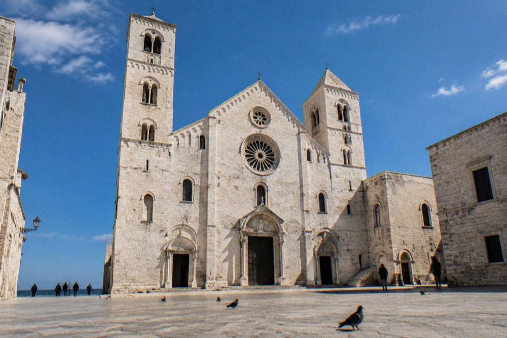 Basilica towers and rose window seen from a low angle across pale paving with pigeons and a deep blue sky