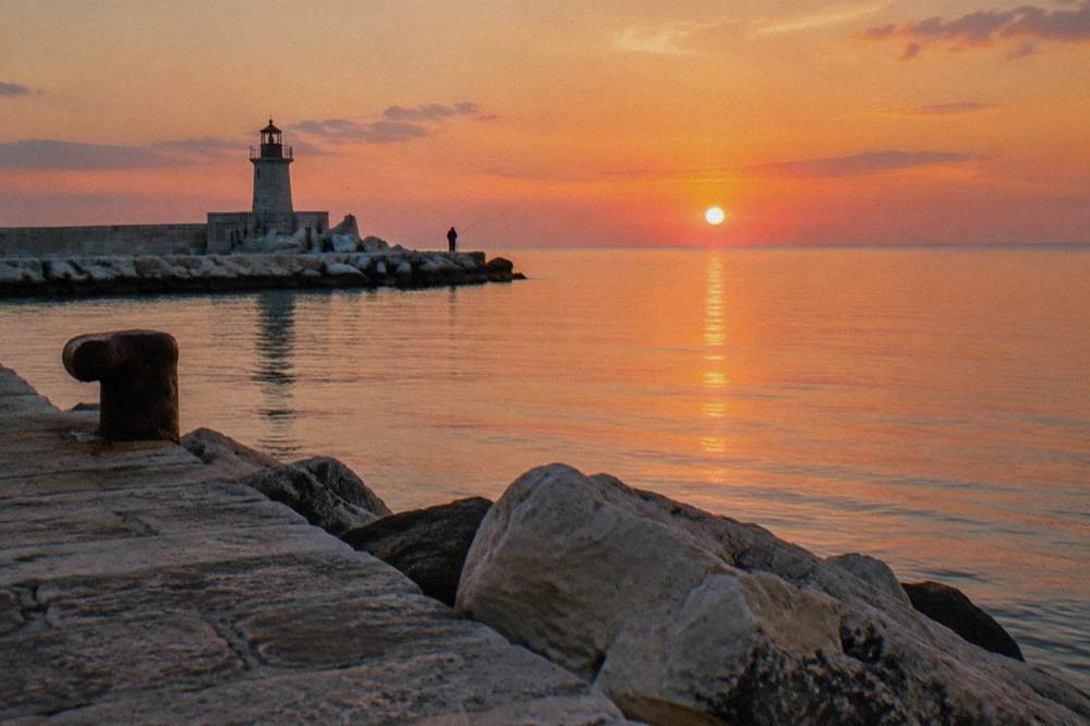 End of a harbour pier with a small lighthouse, glassy open Adriatic and a distant angler at sunset