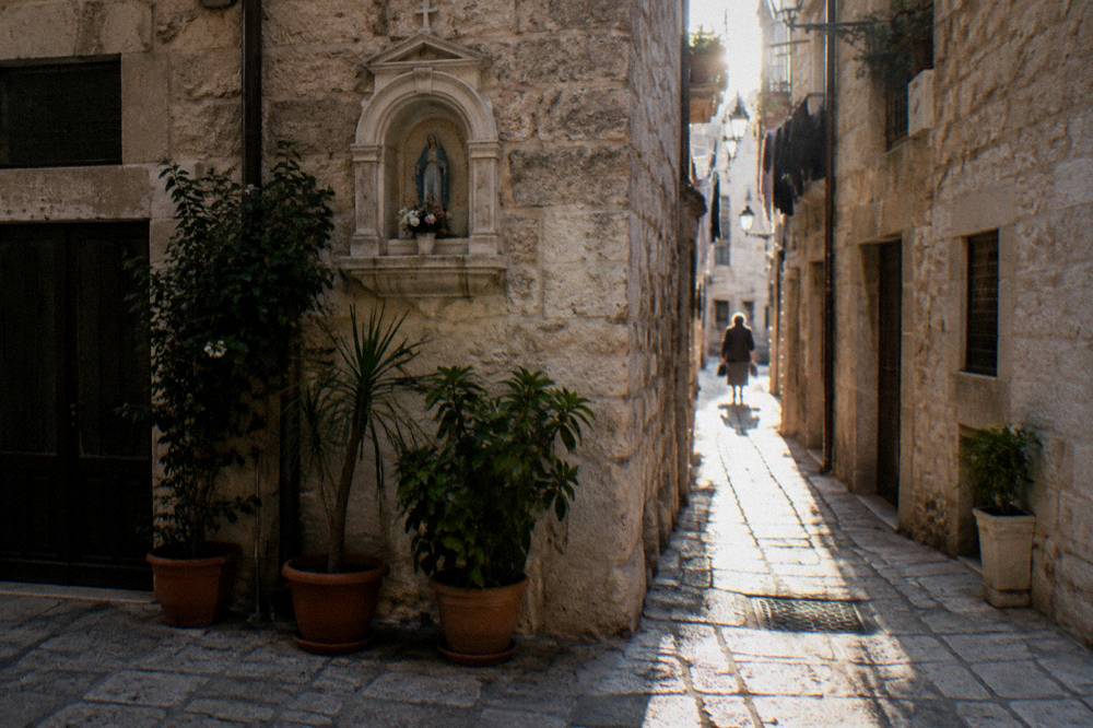 Crossing of narrow old town lanes with a stone shrine niche, potted plants and sunlight slicing between buildings