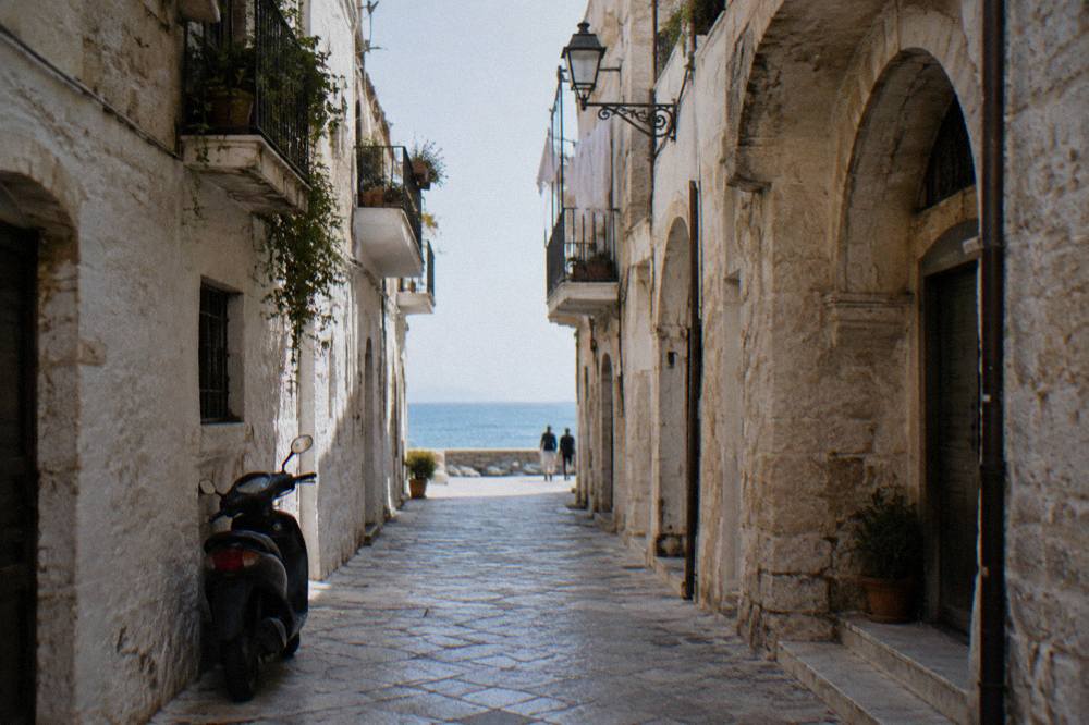 Straight old town lane between whitewashed walls ending in a bright slit of open sea with a parked scooter