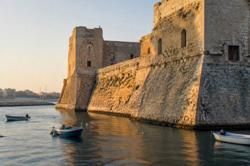 Seaward wall of a pale stone castle beside harbour water with a few small boats in afternoon sun