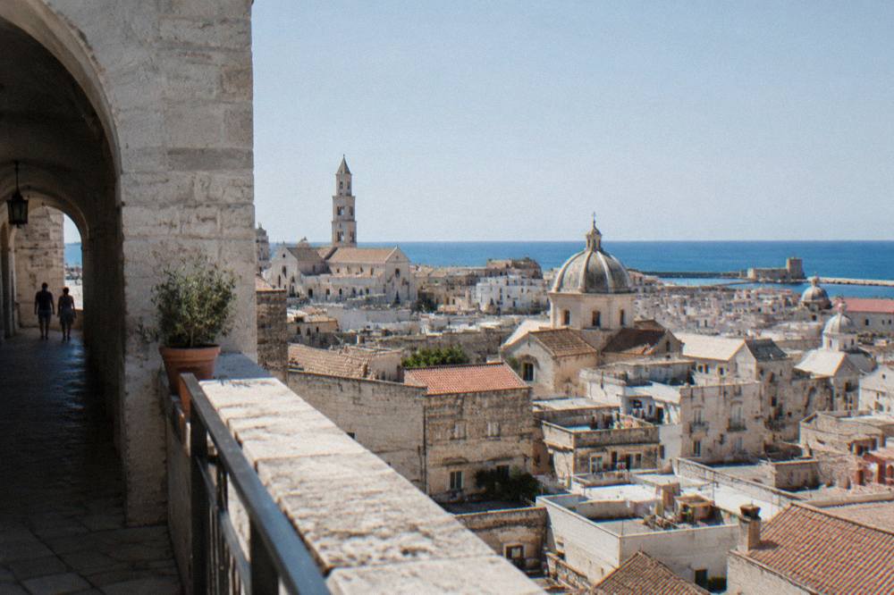Terracotta and limestone rooftops and church domes of a dense old town seen from a high terrace with the sea beyond