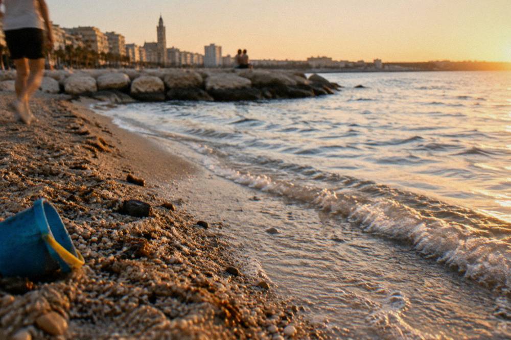 Pebbles and coarse sand at the water's edge with small clear waves and a plastic bucket left on the sand