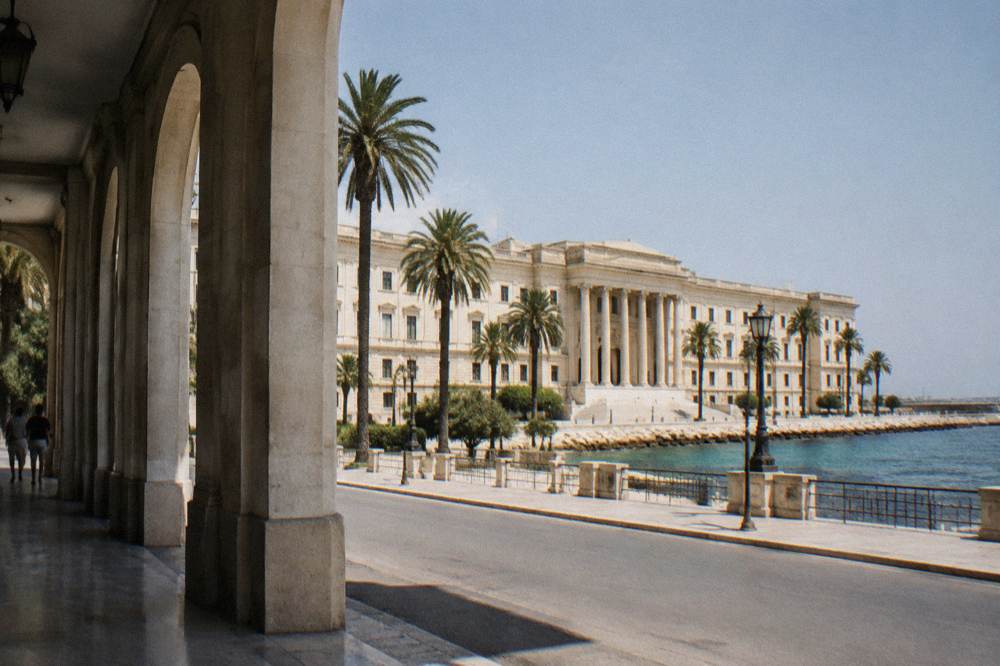 Grand neoclassical seafront palace with a long cream facade, a central portico and palm trees