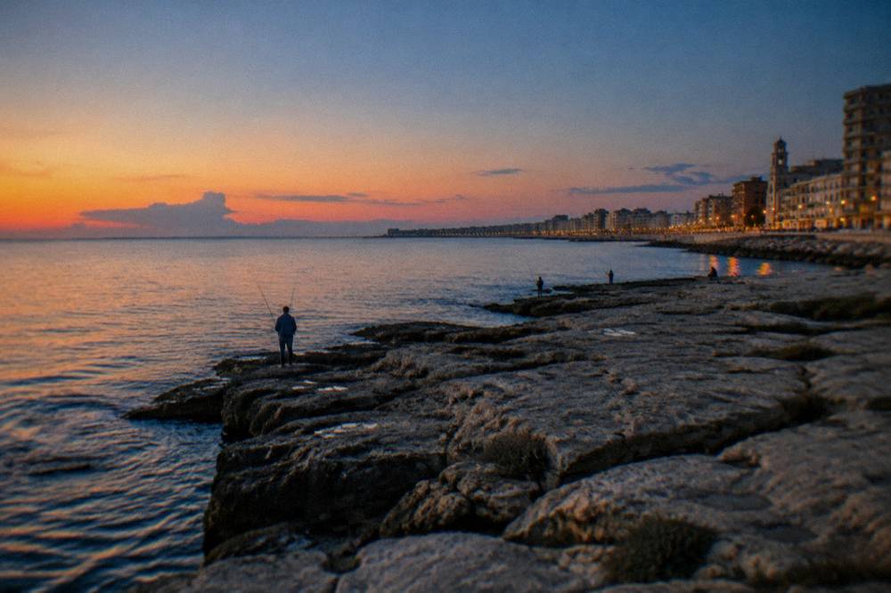 Seafront limestone rocks below a residential district with distant anglers and calm water in early evening