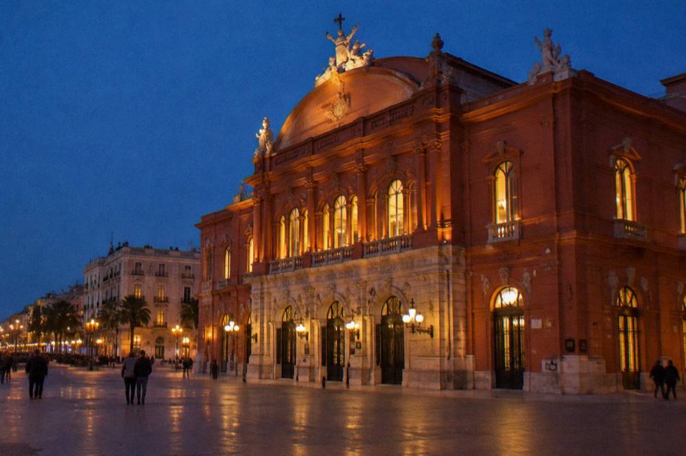 Neoclassical theatre facade floodlit at evening with glowing arched windows and a deep blue sky