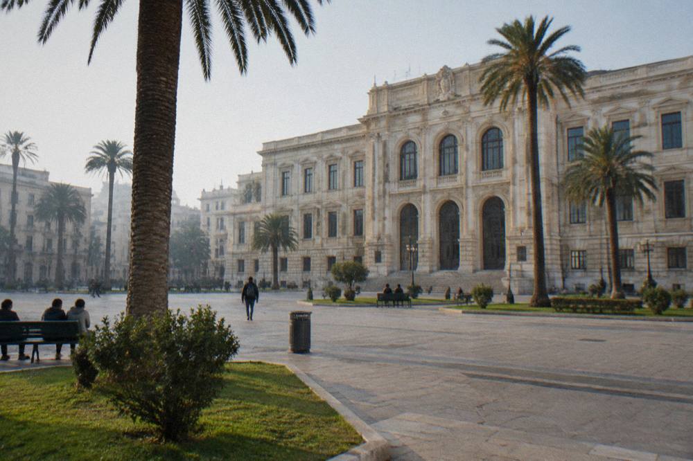 Broad city square in front of a monumental cream stone university building with palm trees, lawns and benches