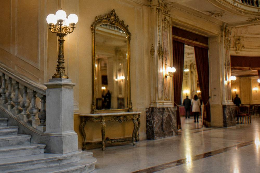 Quiet gilded theatre foyer with a marble staircase and a tall mirror in soft warm light