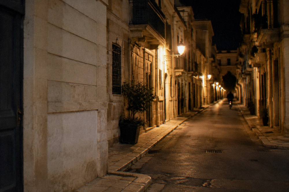 Quiet side street at night with warm lamps, a distant cyclist and pale stucco facades