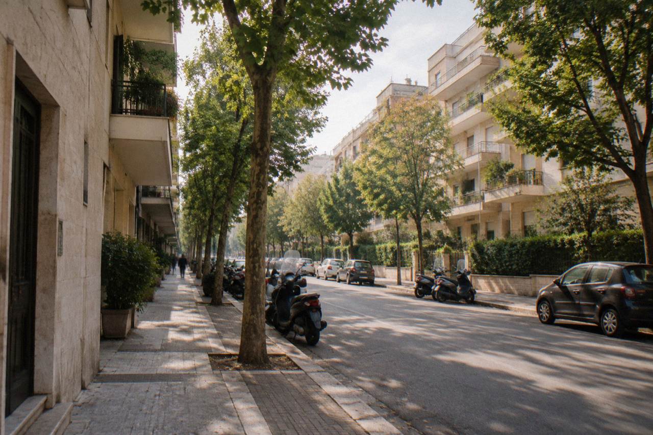Leafy residential avenue of mid-century apartment blocks with street trees and parked scooters