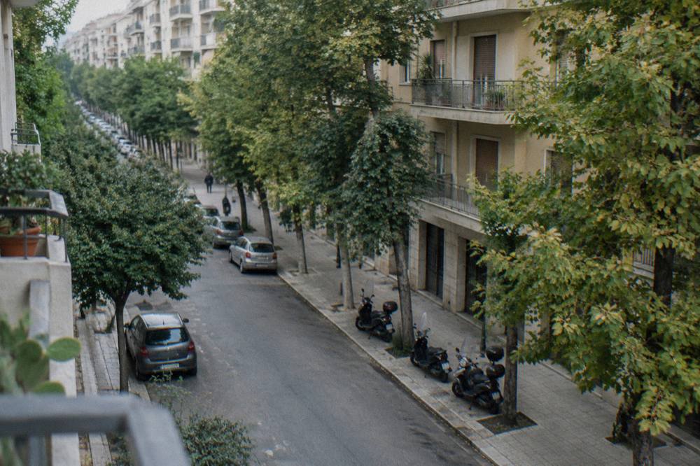 Leafy residential avenue of mid-century apartment blocks with street trees and parked scooters