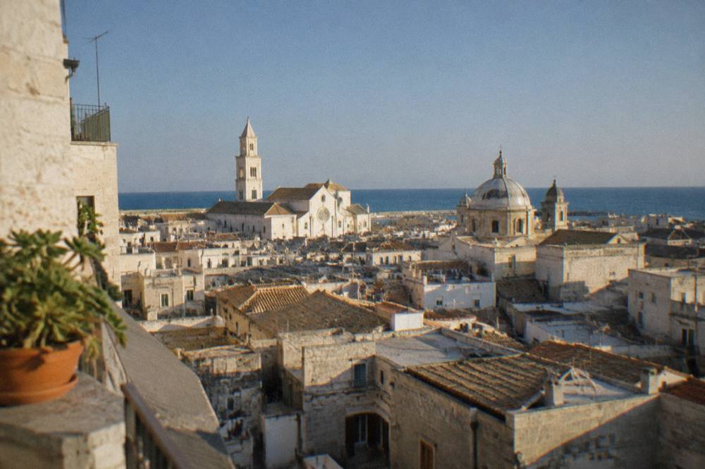Terracotta and limestone rooftops and church domes of a dense old town seen from a high terrace with the sea beyond