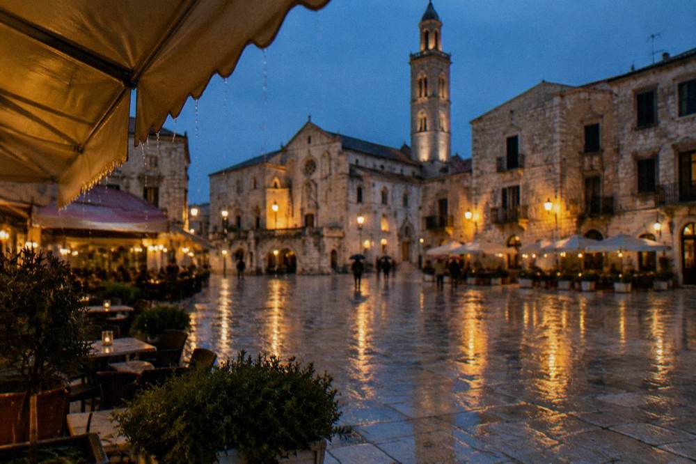 Rainy evening in a pale stone square with wet paving reflecting warm cafe lights and distant umbrellas