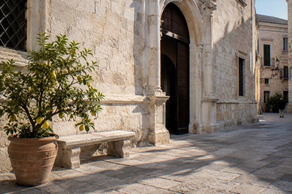 Pale stone palazzo with an arched portal on a quiet old-town street beside a stone bench and a potted lemon tree