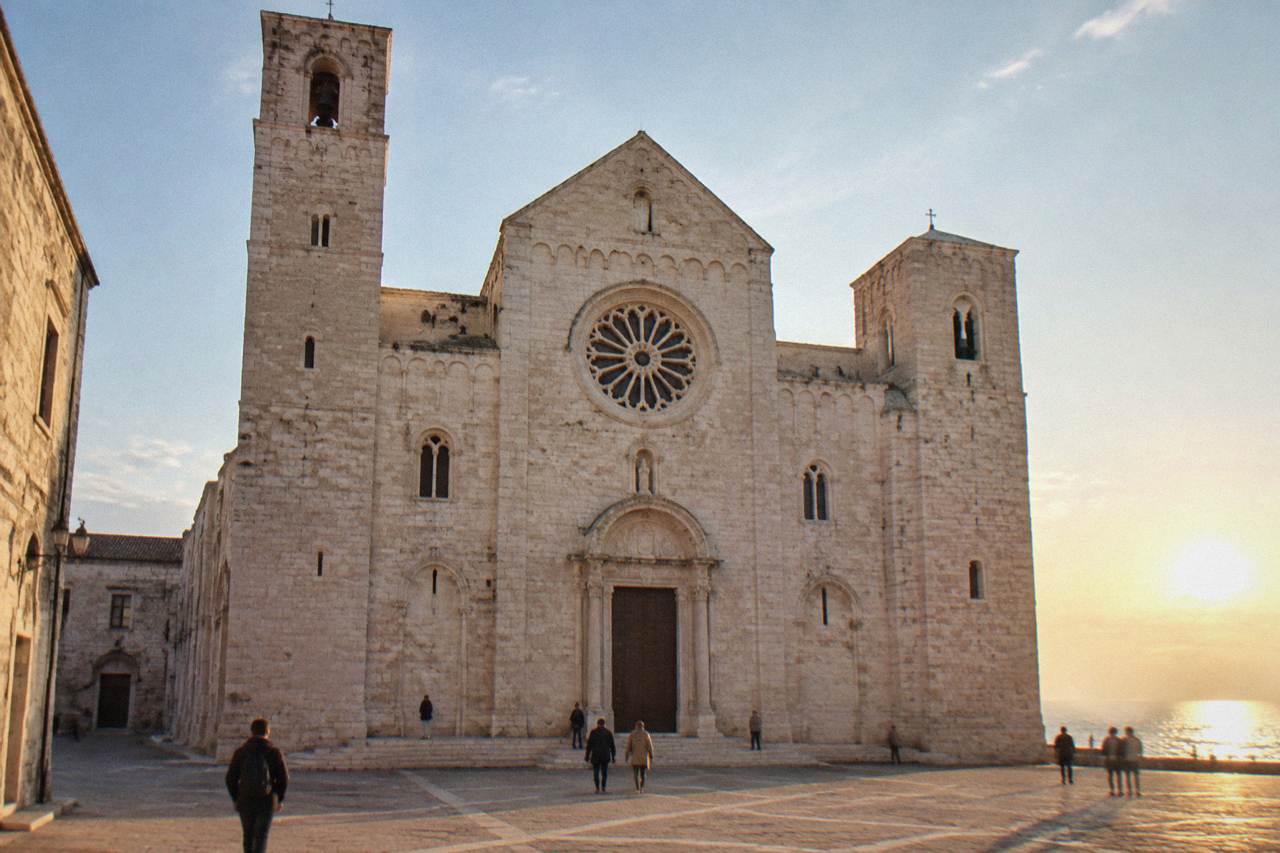 Romanesque church of pale limestone with a wide plain facade, two squat towers and a rose window above a paved square