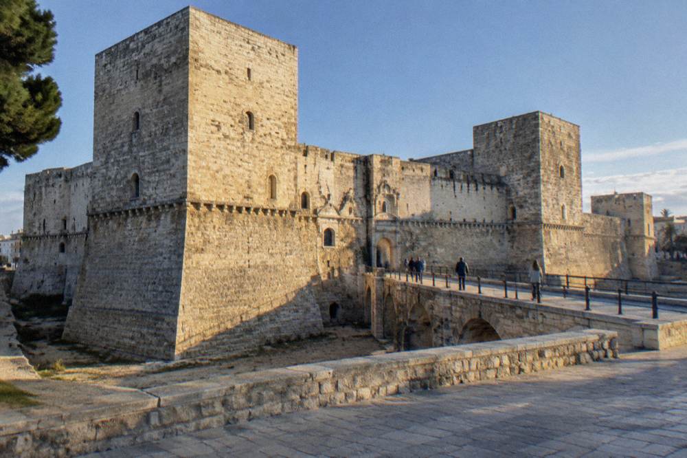Massive square pale limestone fortress with corner towers, a dry moat and a bridge leading to an arched gate