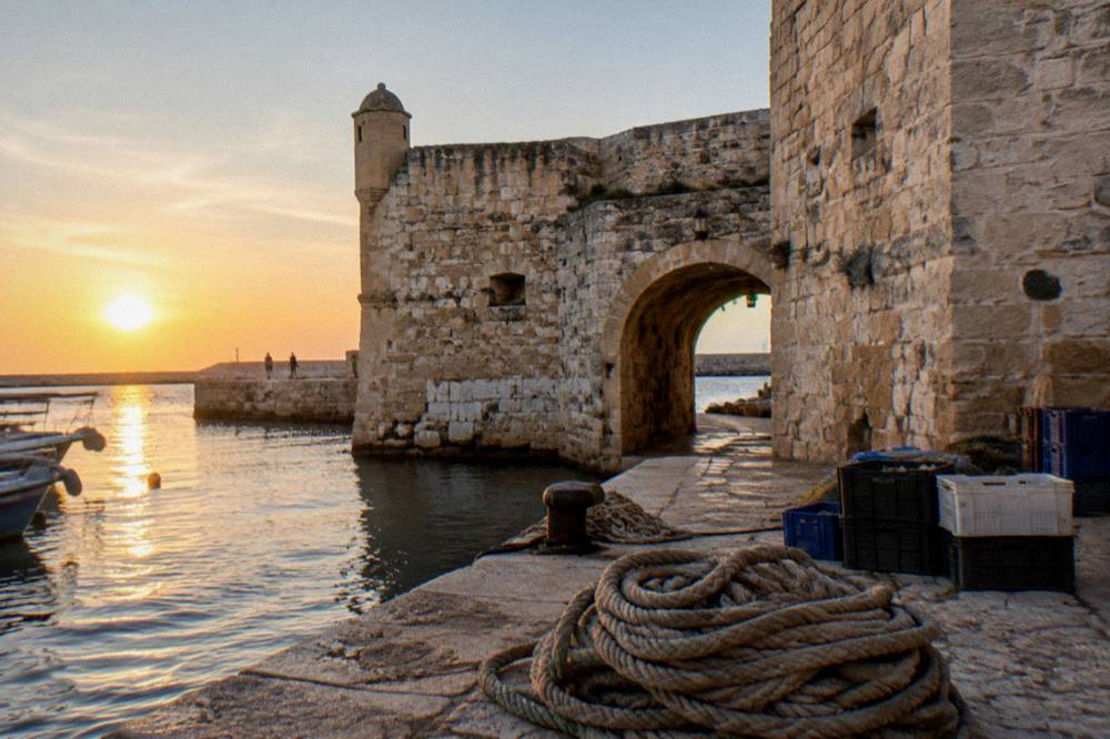 Weathered limestone fort walls and a low arched gateway beside harbour water with coiled ropes and crates on the quay