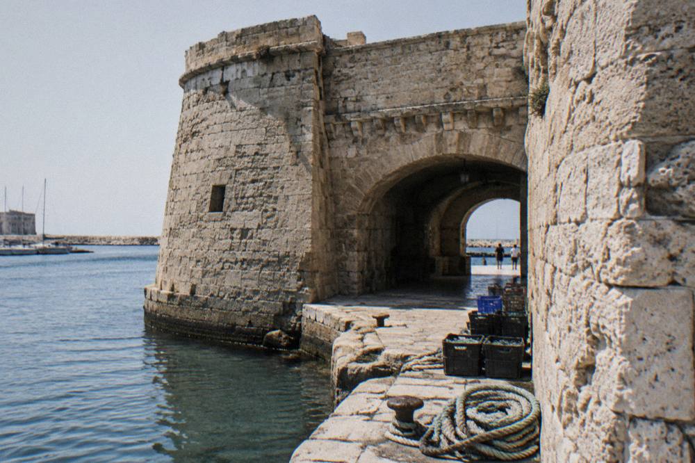 Weathered limestone fort walls and a low arched gateway beside harbour water with coiled ropes and crates on the quay