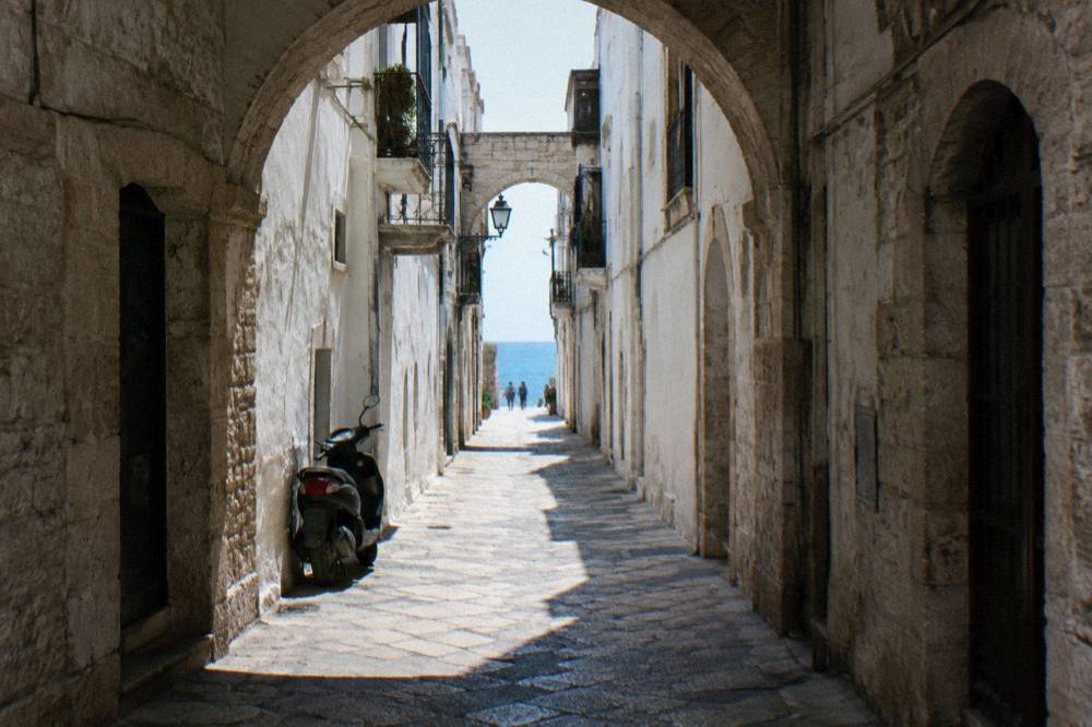 Straight old town lane between whitewashed walls ending in a bright slit of open sea with a parked scooter