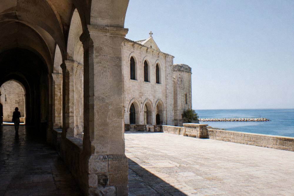 Low pale stone former monastery beside the sea with arched windows and a bare stone terrace