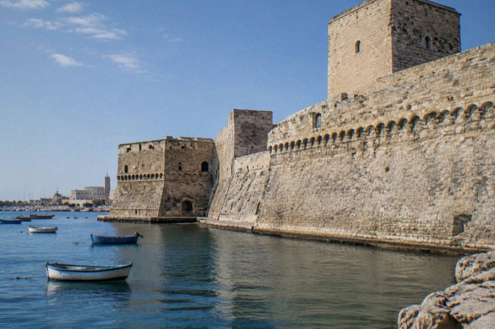 Seaward wall of a pale stone castle beside harbour water with a few small boats in afternoon sun
