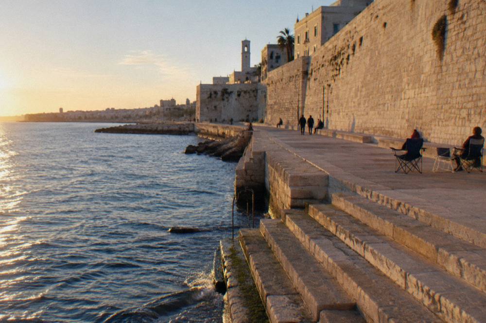Massive pale limestone sea wall with a promenade at its foot and worn steps down to the water in low afternoon sun