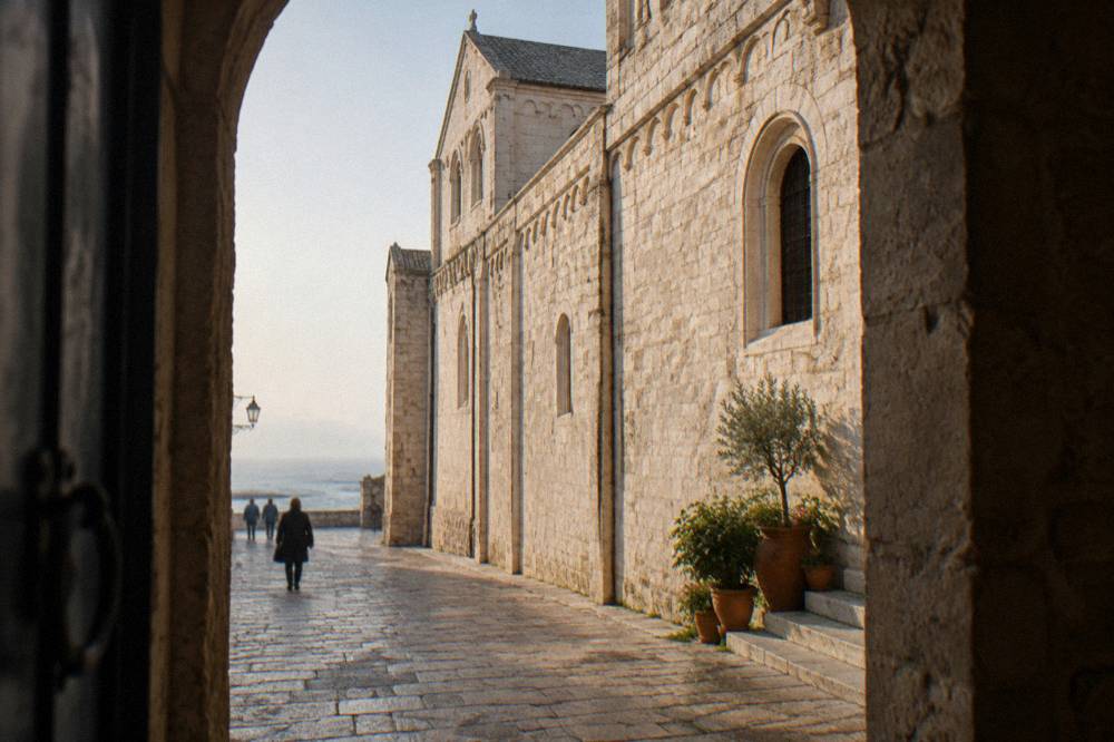 Long pale limestone flank of a Romanesque church seen from a narrow old-town lane with pots on a doorstep