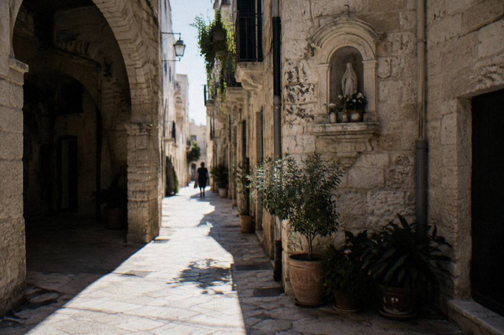 Crossing of narrow old town lanes with a stone shrine niche, potted plants and sunlight slicing between buildings