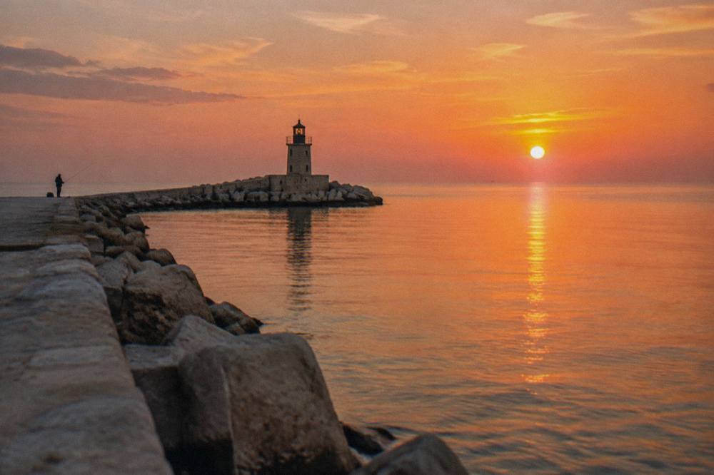 End of a harbour pier with a small lighthouse, glassy open Adriatic and a distant angler at sunset