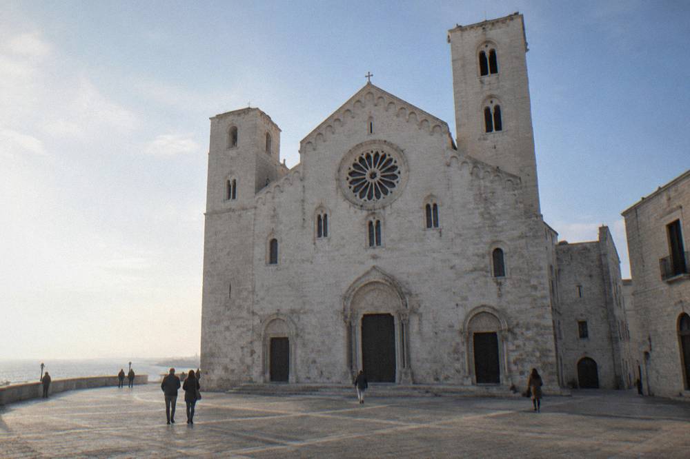 Romanesque church of pale limestone with a wide plain facade, two squat towers and a rose window above a paved square