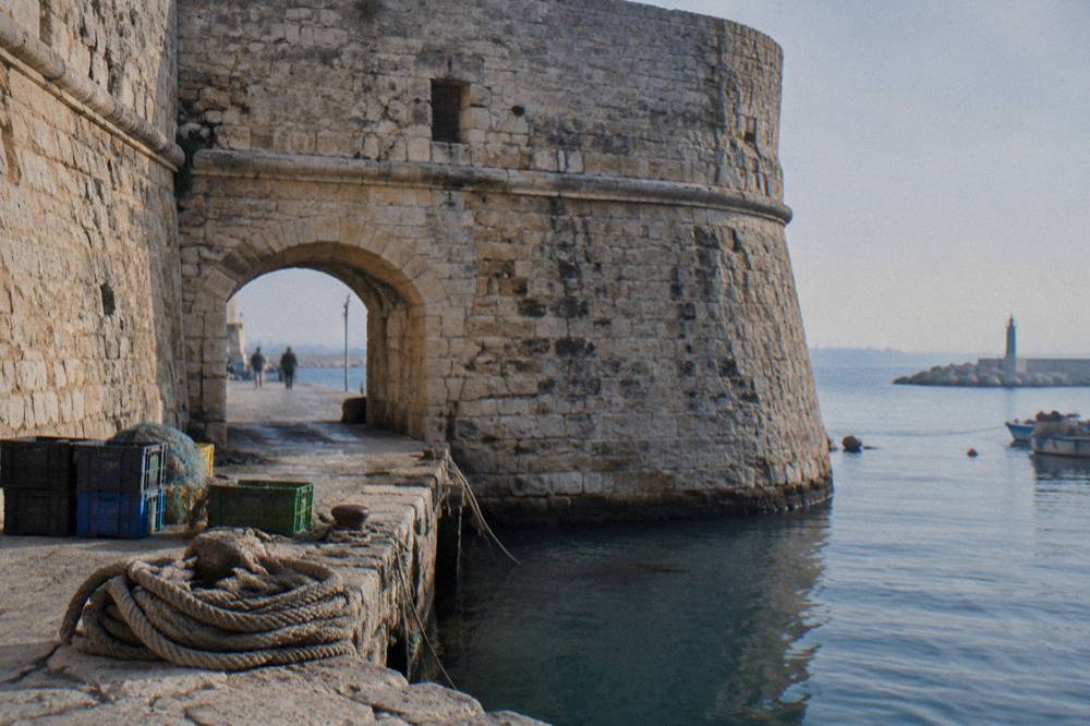 Weathered limestone fort walls and a low arched gateway beside harbour water with coiled ropes and crates on the quay