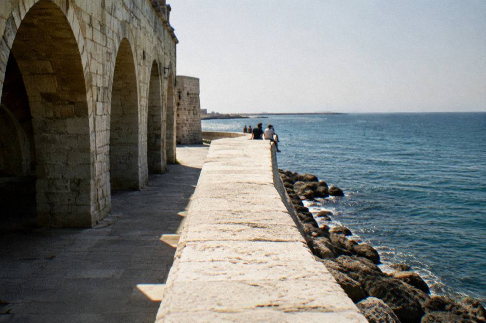 Distant people sitting on the broad top of a pale limestone sea wall above the open Adriatic in soft warm light