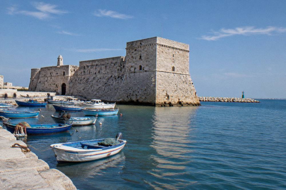 Squat pale limestone fort with thick sloping walls at the edge of the old harbour with small fishing boats moored in front