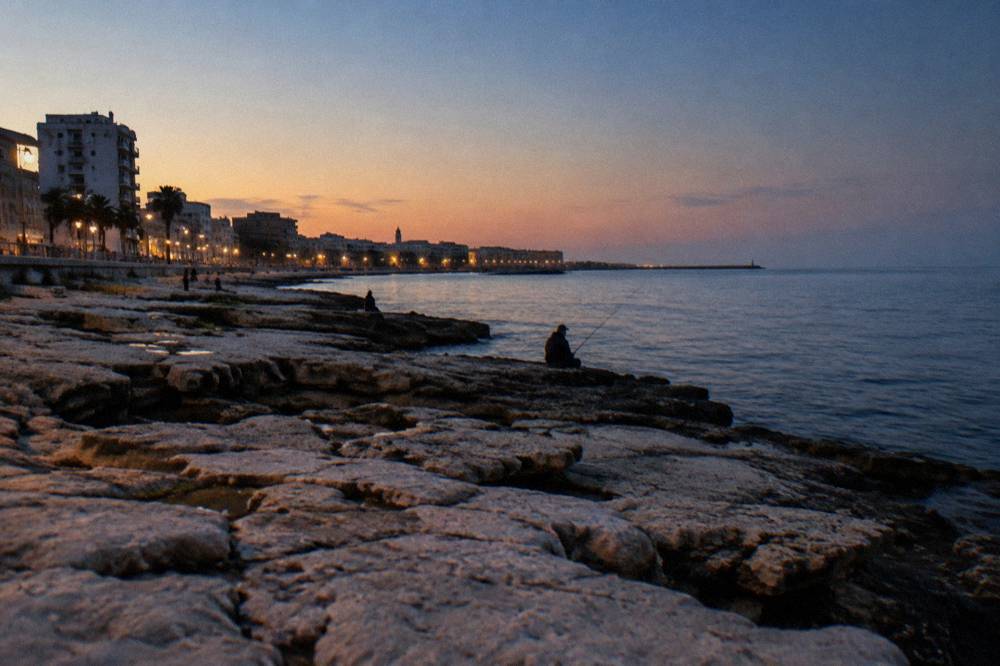 Seafront limestone rocks below a residential district with distant anglers and calm water in early evening