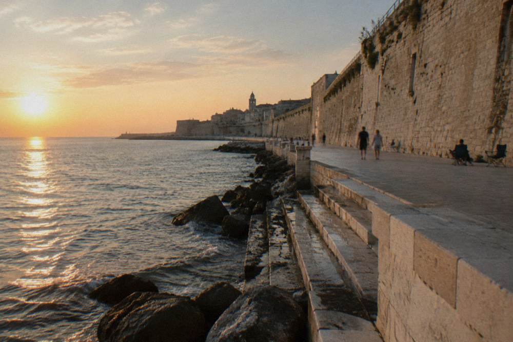 Massive pale limestone sea wall with a promenade at its foot and worn steps down to the water in low afternoon sun