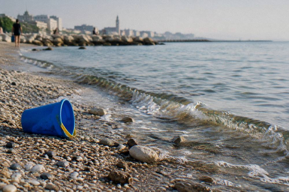 Pebbles and coarse sand at the water's edge with small clear waves and a plastic bucket left on the sand